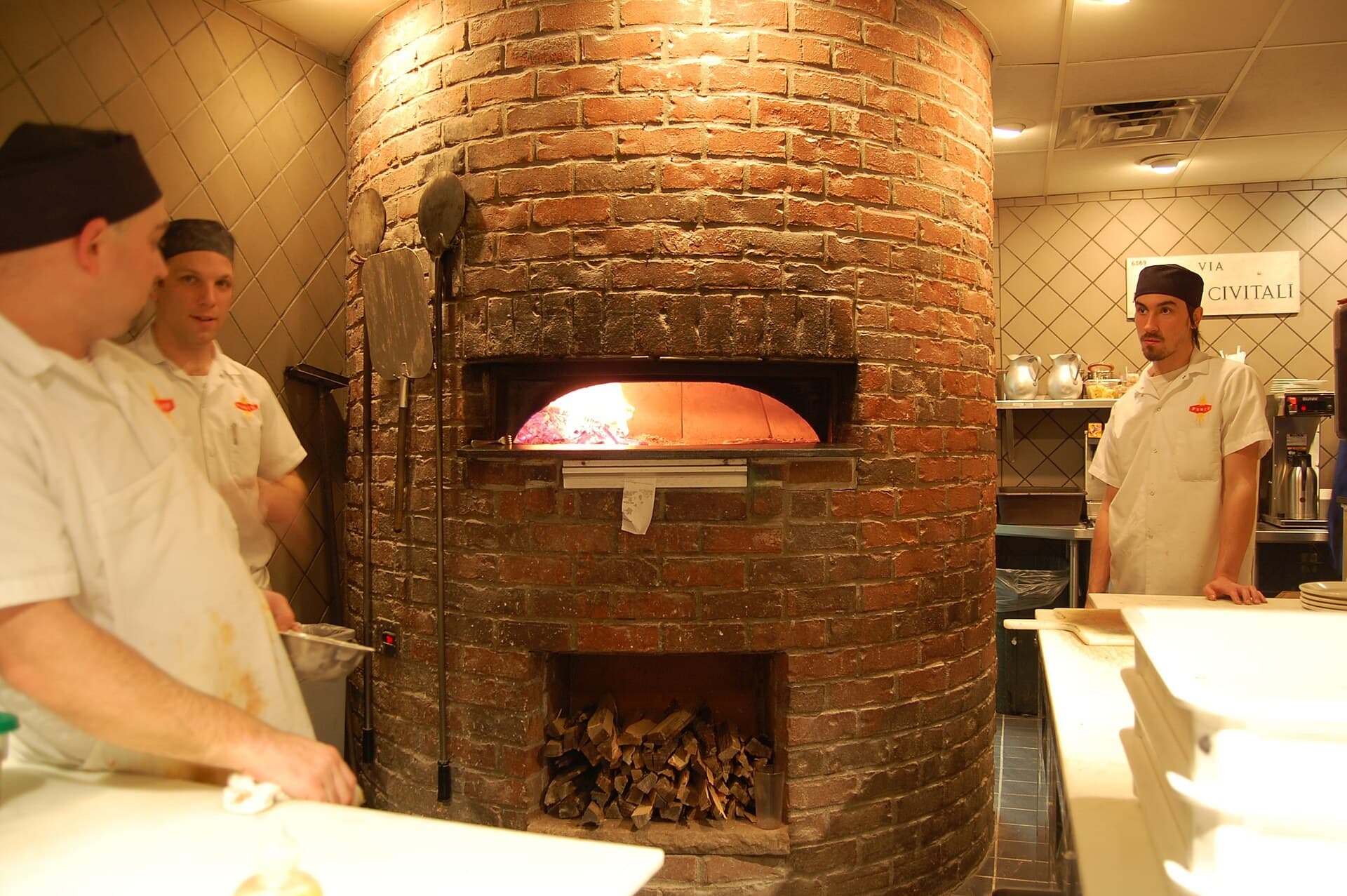 Kitchen staff beside a round brick pizza oven in a pizzeria