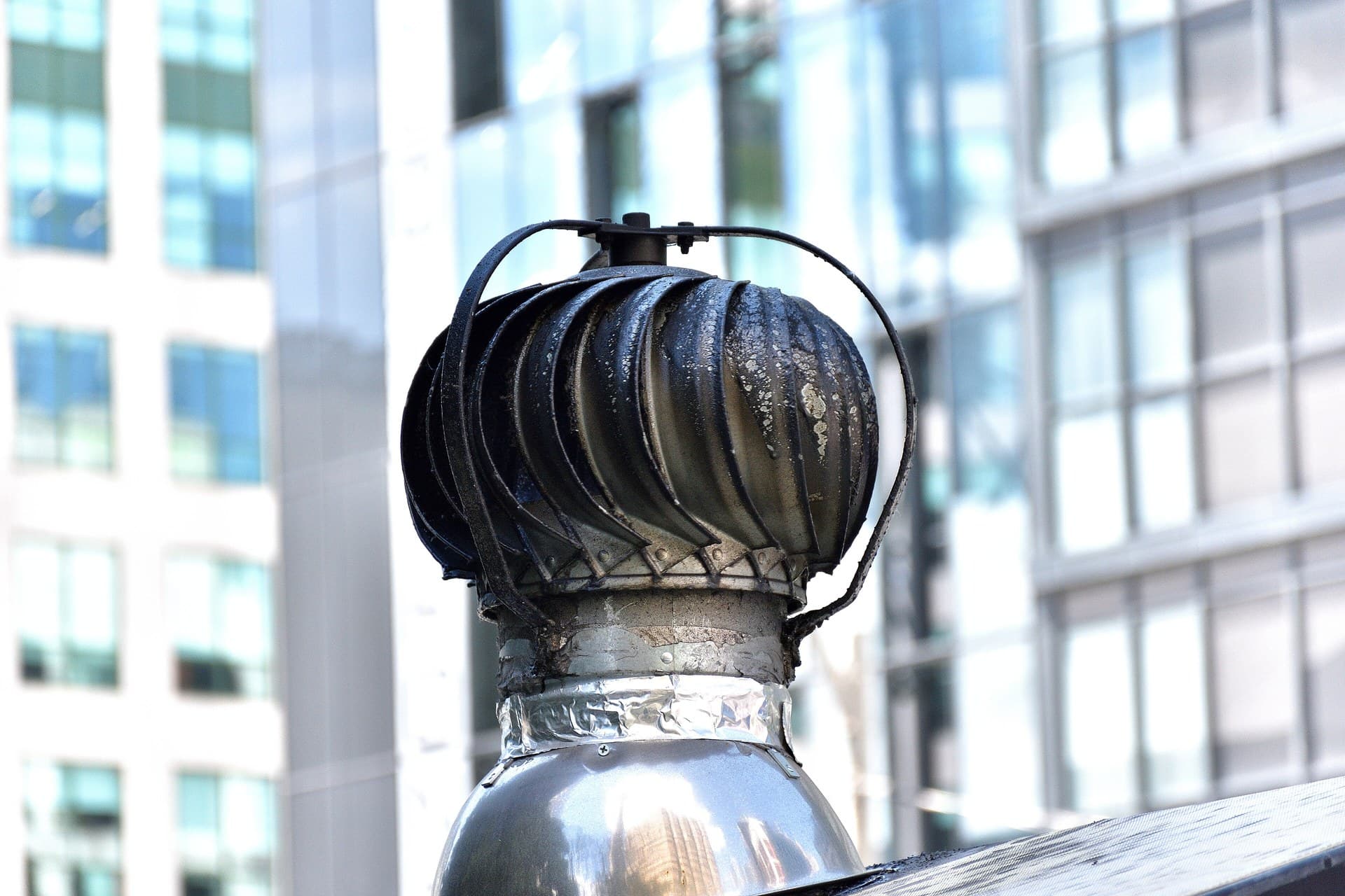 A weathered roof cyclone (turbine ventilator) with curved vanes on its throat, against city buildings